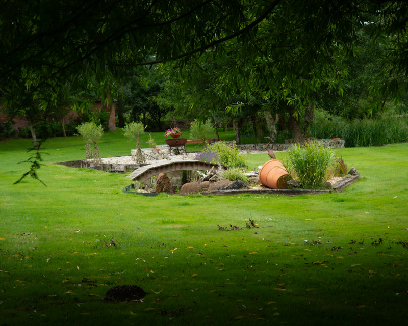 Tranquil green lawn with a small stone bridge crossing a pond, an upturned terracotta pot, and lush foliage