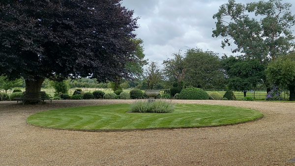 Neatly mowed circular lawn with striped pattern, surrounded by topiary shrubs and a grand tree